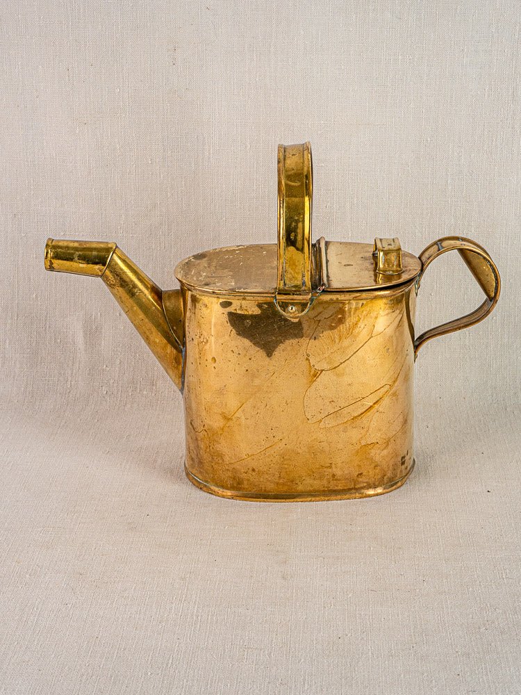 Antique French brass watering can circa 1900 with hinged lid and dual handles on fabric background.