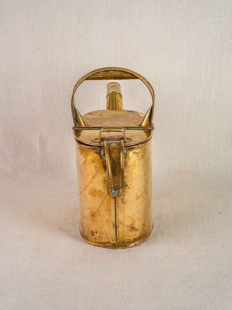 Antique French brass watering can with hinged lid and top handle on light fabric background.