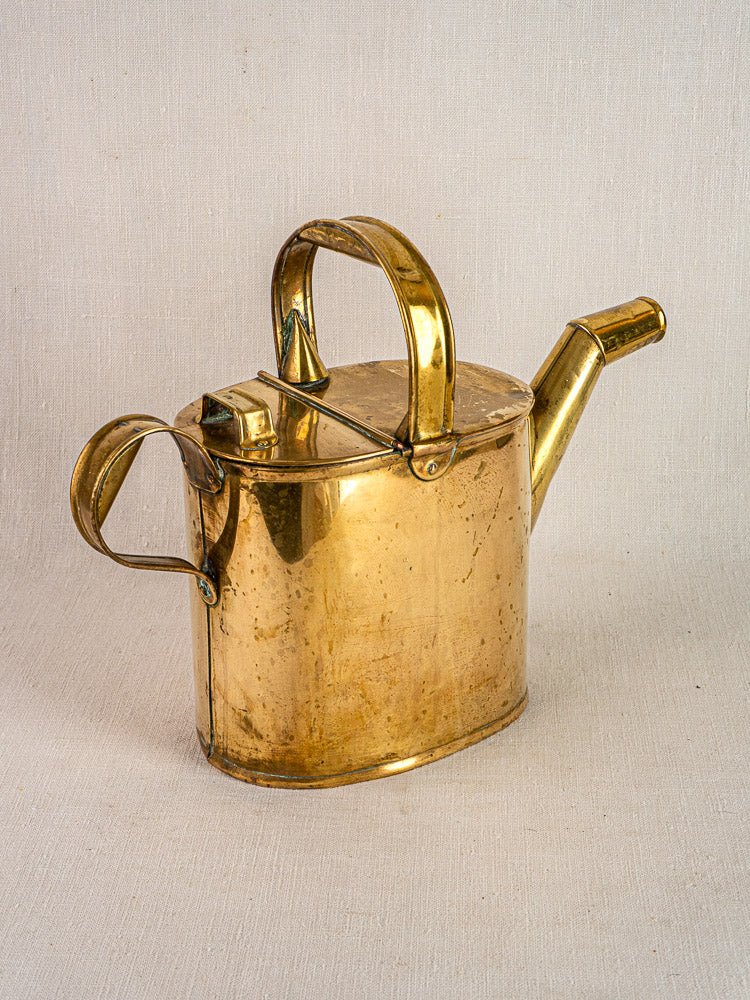 Antique French brass watering can with hinged lid, double handles, and long spout on fabric.