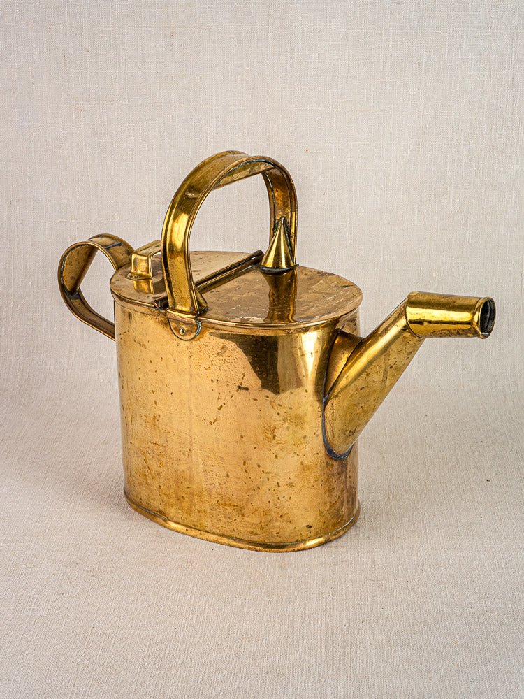 Antique French brass watering can circa 1900 with rounded handle, curved spout, and hinged lid on neutral fabric.