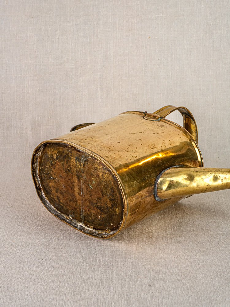 Antique French brass watering can with hinged lid, curved handle, and aged patina on fabric.