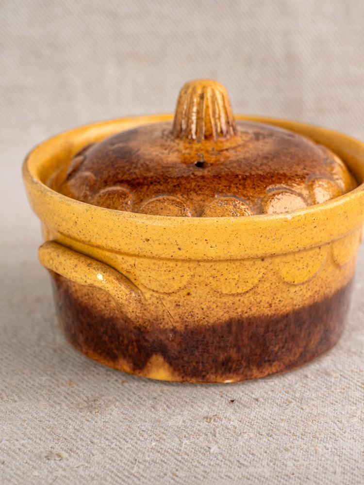 Antique French Alsace earthenware cocotte with lid, small handle, and decorative lid patterns on light textured fabric.