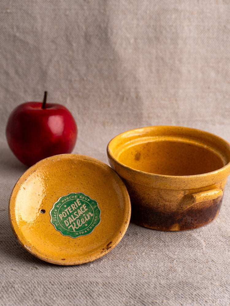 Antique French Klein Strasbourg yellow-brown glazed earthenware cocotte with lid removed, beside red apple on beige fabric.