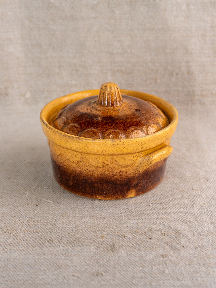 Antique French earthenware cocotte from Klein Strasbourg with lid in rustic brown and yellow, on beige fabric.