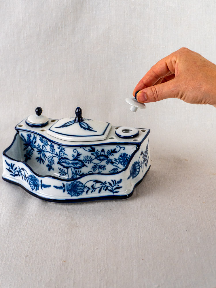 Blue and white porcelain inkstand with floral motifs, lid being lifted, on light fabric background.