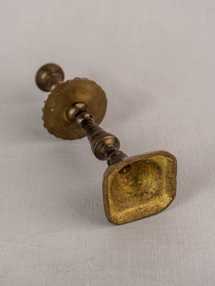 Antique French brass candlestick with scalloped drip pan, worn golden square base on light fabric.