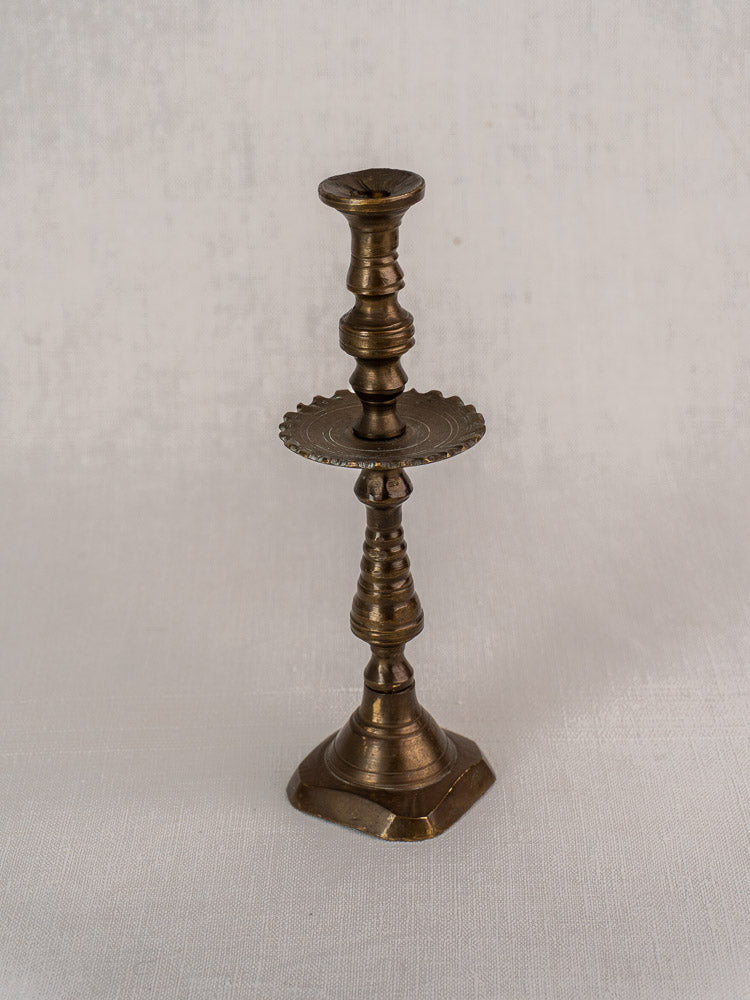 Antique French brass candlestick with scalloped drip pan and square base on textured white surface.