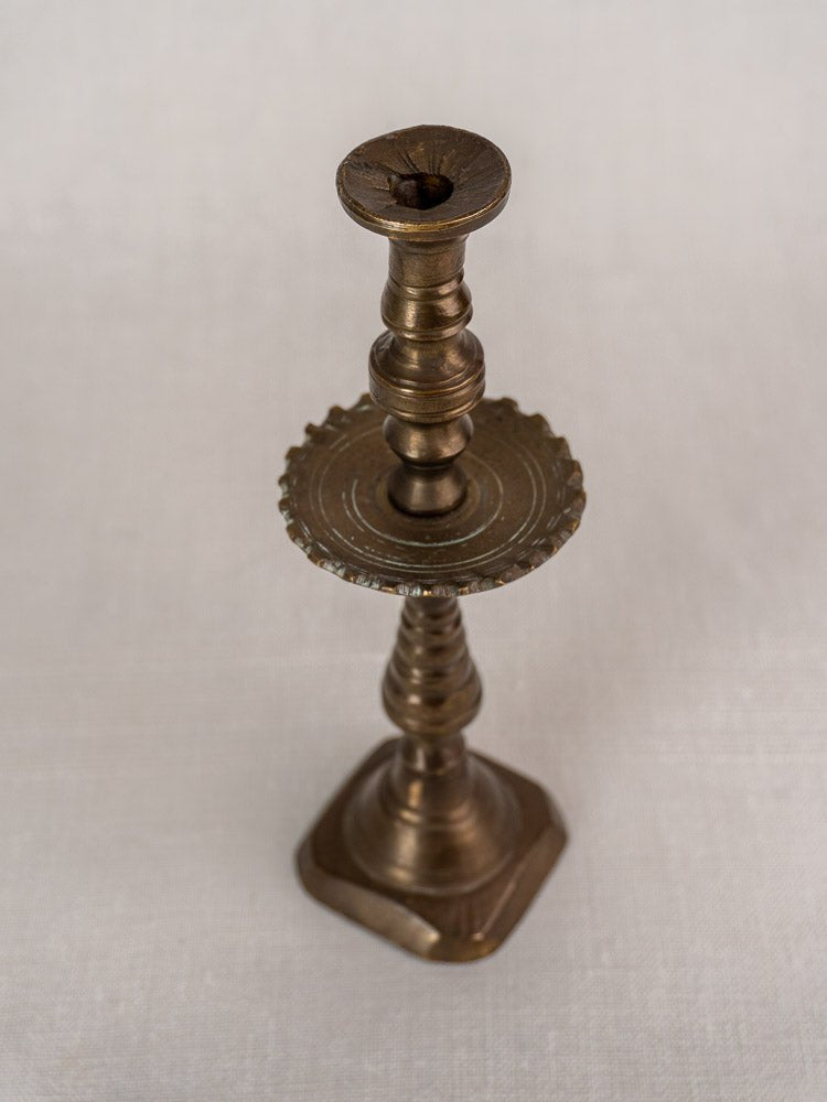 19th-century French brass candlestick with scalloped drip pan and square base on neutral fabric.