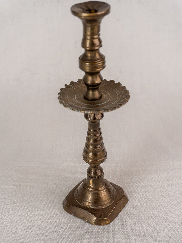 19th century French brass candlestick with scalloped drip pan and square base on textured surface.