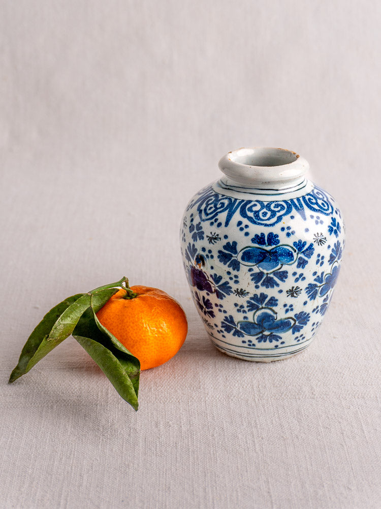 Antique Dutch Delft blue and white faience vase with chinoiserie florals beside a mandarin orange on light cloth.