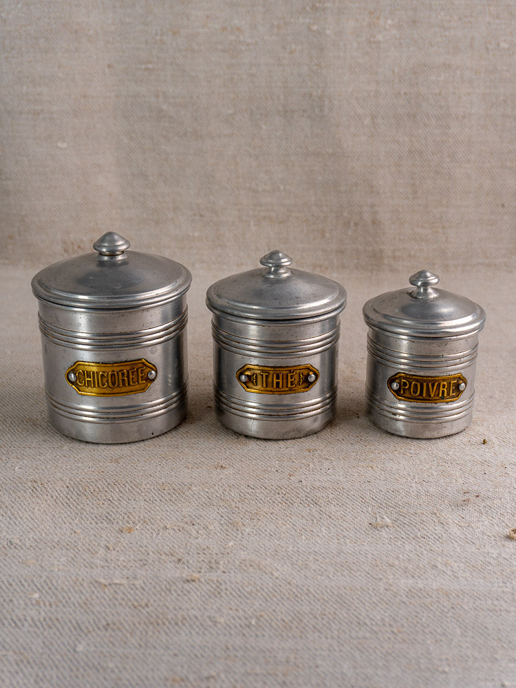 Vintage French aluminum canister set with brass labels Chicorée, Thé, Poivre; matching lids on beige textured fabric.