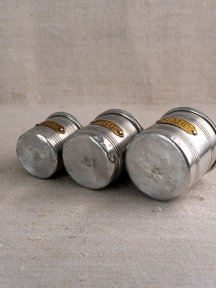 Vintage French aluminum canister set with brass labels and lids, mid-century, on textured beige fabric, worn scuffs visible.