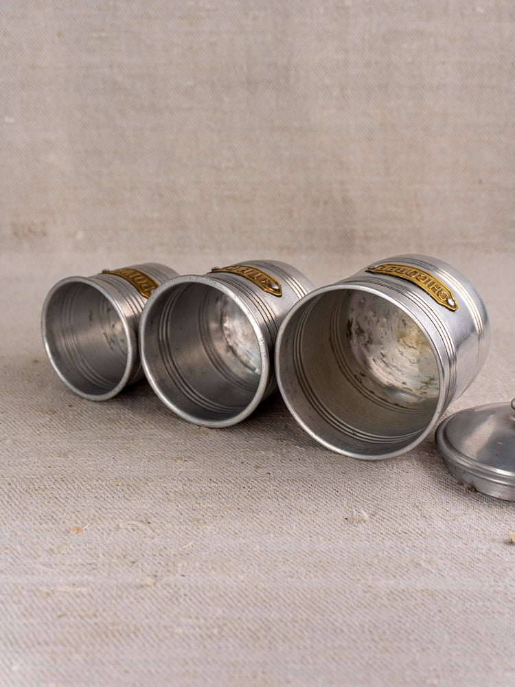 Mid-century French aluminum canister set with brass labels, three pieces and a lid, displayed on beige fabric.
