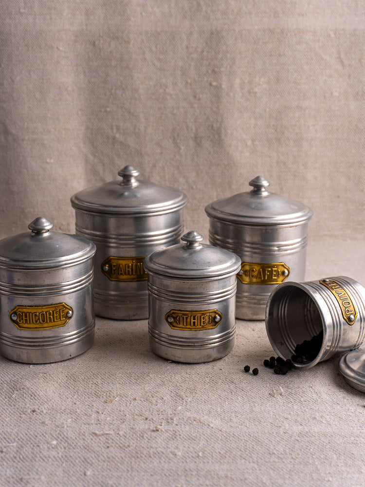 Vintage French aluminum canister set with brass labels, five lidded pieces displayed on textured beige fabric.
