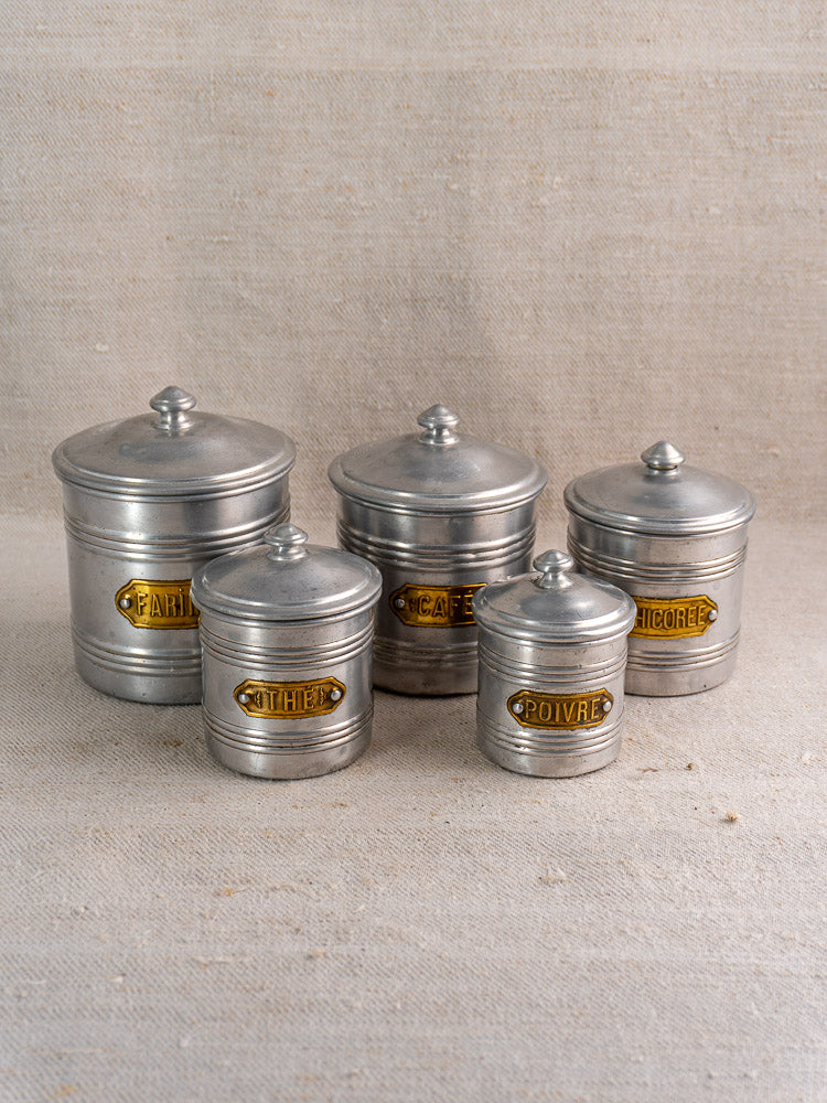 Vintage French aluminum canister set with brass labels, five lidded pieces displayed on textured beige fabric.