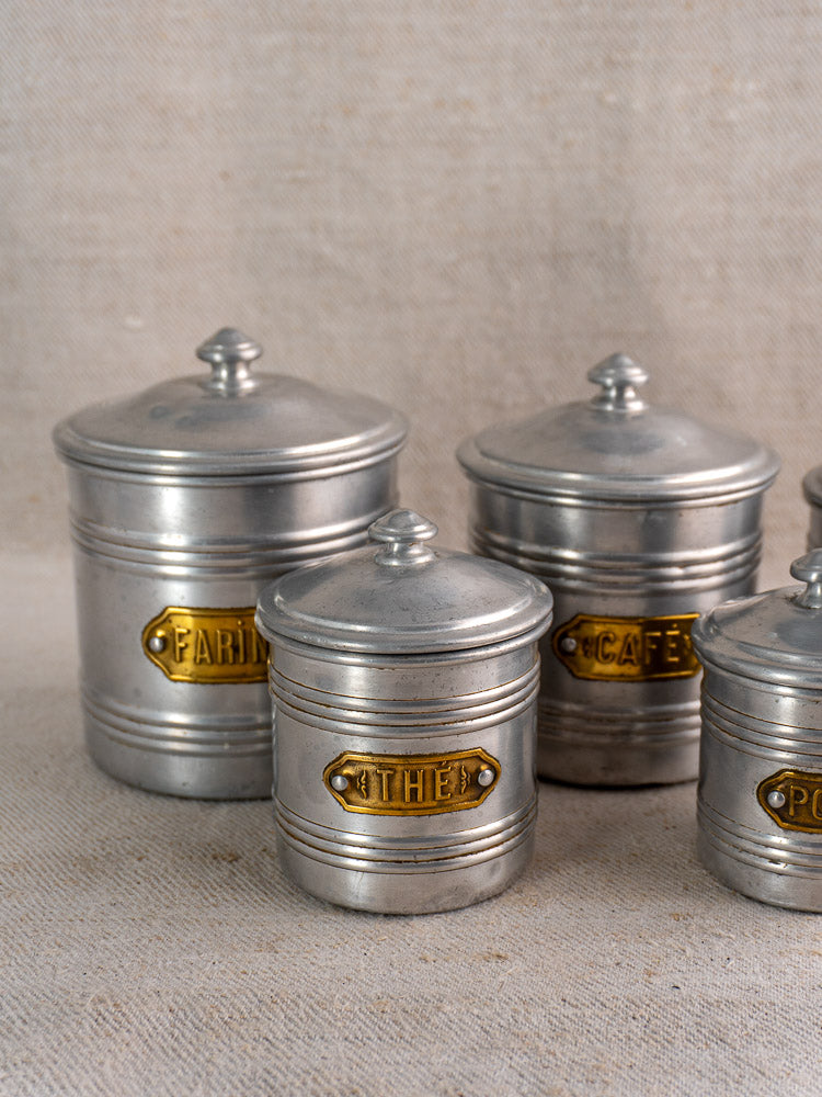 Vintage French aluminum canister set with brass labels for farine, thé, café, and poivre on beige fabric.