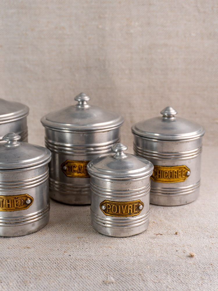 Mid-century French aluminum canister set with brass labels, five lidded pieces, gently worn, on neutral fabric.