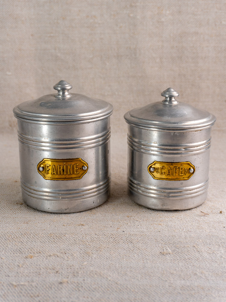 Pair of vintage mid-century French aluminum canisters with brass FARINE and CAFÉ labels on neutral fabric.