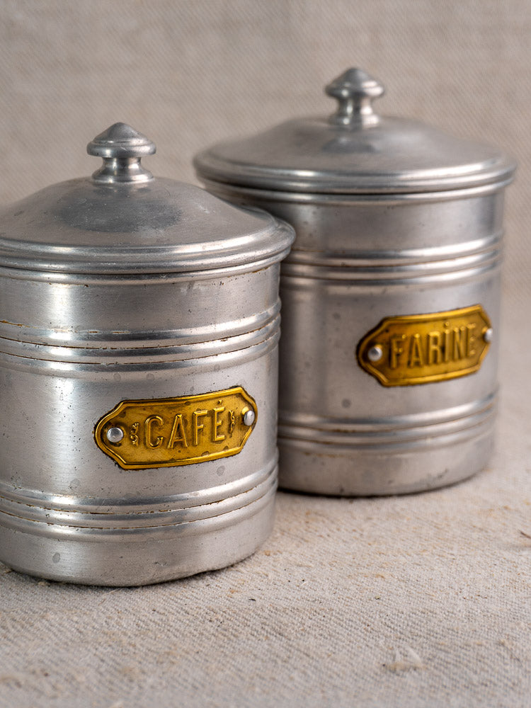 Vintage French aluminum canister set with mid-century brass “CAFÉ” and “FARINE” labels on neutral fabric surface.