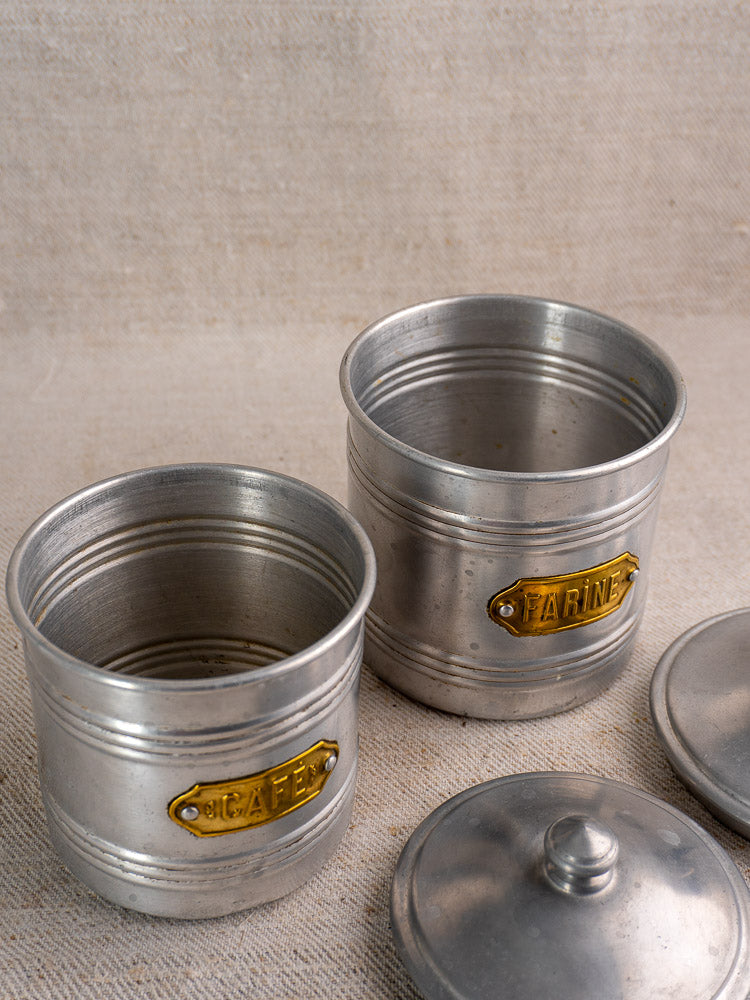 Mid-century French aluminum canister set with brass labels, CAFÉ and FARINE, on beige fabric surface, lids displayed alongside.