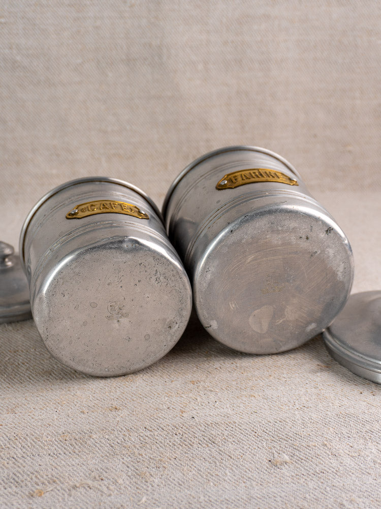 Mid-century French aluminum canister set with brass labels, on textured beige fabric, visible wear and lid nearby.