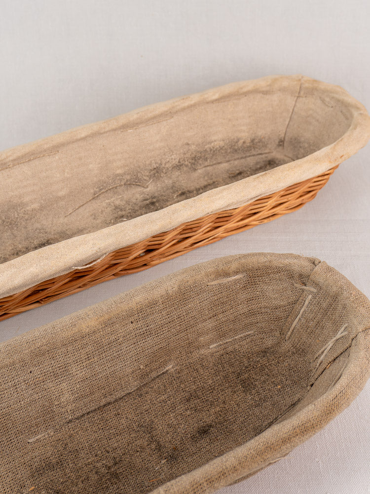 Pair of vintage French baguette proofing baskets with linen liners showing flour marks and creases on a pale surface.