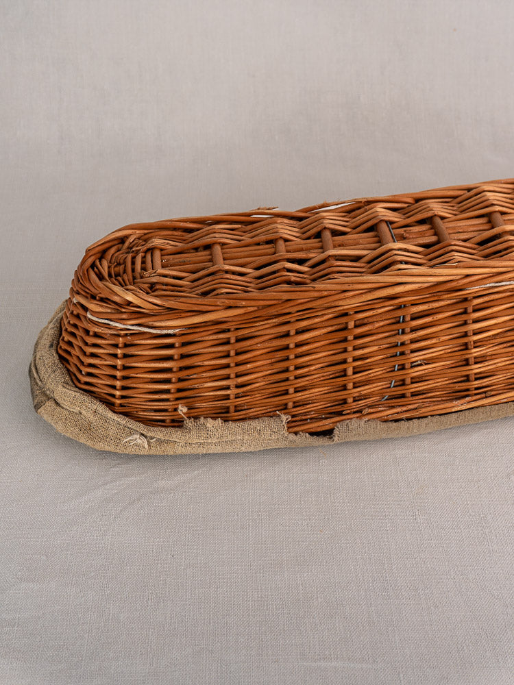 Vintage French baguette proofing basket, long oval shape with linen liner, on light gray fabric surface.