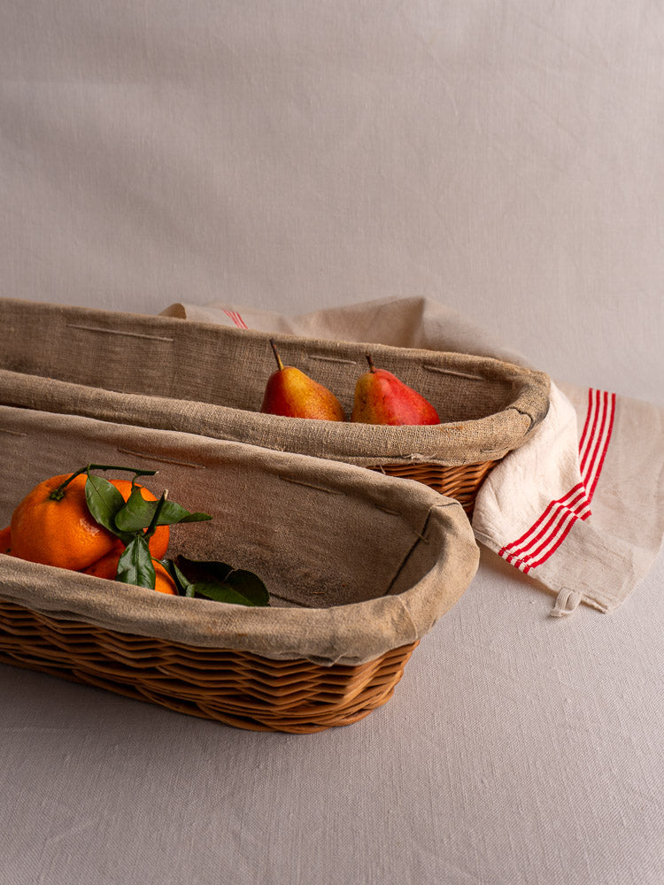 Pair of vintage French baguette proofing baskets with linen liners, styled with fruit and cream-striped cloth.