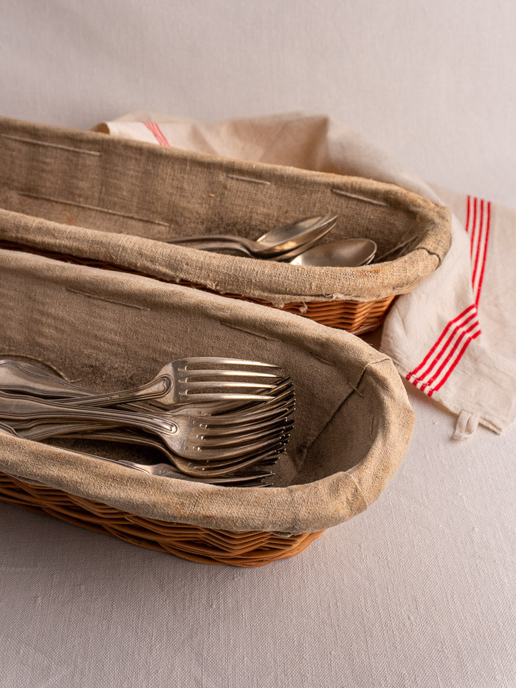 Pair of vintage French baguette proofing baskets with linen liners, filled with metal flatware, beside red-striped white cloth.