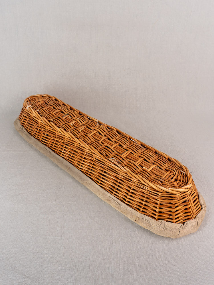Vintage French wicker baguette proofing basket with linen liner, circa 1940-1950, on a pale fabric background.