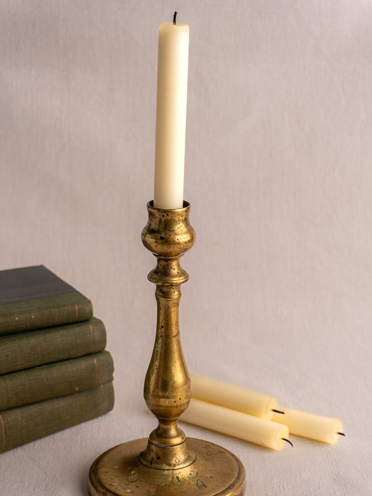 Antique French brass candlestick with white candle, alongside books and candles on neutral fabric.