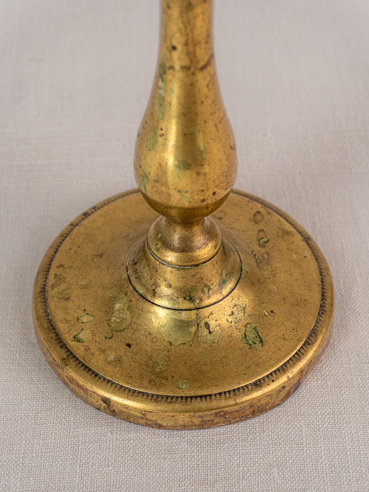 Antique French brass candlestick base with baluster stem, worn finish, patina, and engraved details on beige surface.
