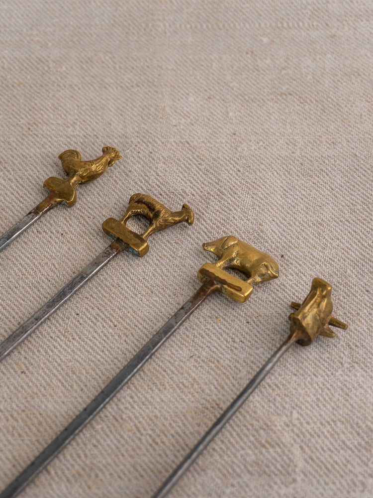 Set of four vintage French brochette skewers with brass animal finials and wooden stand on beige fabric.