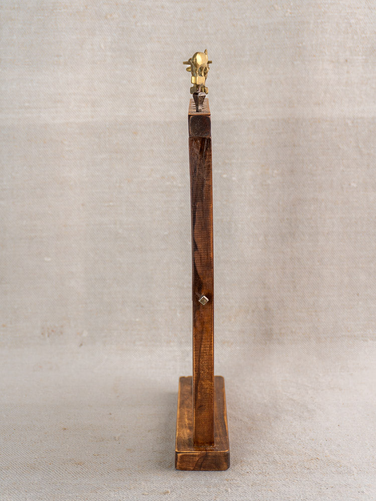 Tall wooden sculpture with petite brass animal finial, displayed against a beige fabric backdrop.