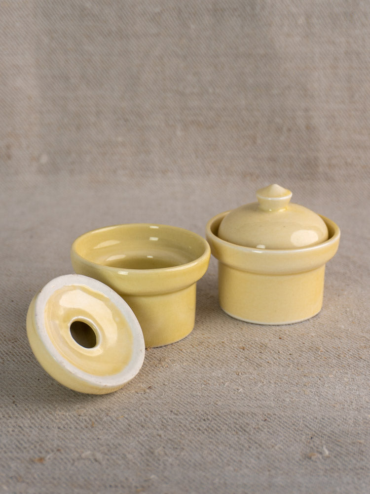 Pair of mid-20th century French cream glazed butter pots with unglazed bases on a beige surface, one lidded, one open.