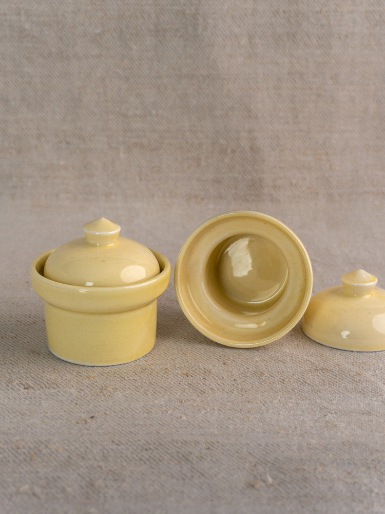 Pair of mid-century cream-glazed French beurriers, one open, one closed, on textured beige fabric.