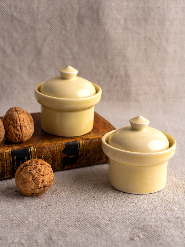 Pair of vintage French cream-glazed butter pots with lids, one on a brown book, three walnuts nearby on beige textured surface.