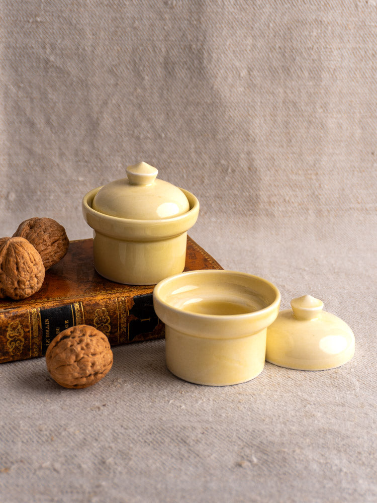 Pair of vintage French butter pots with cream glaze, one open, one closed on a book, with walnuts on beige fabric.