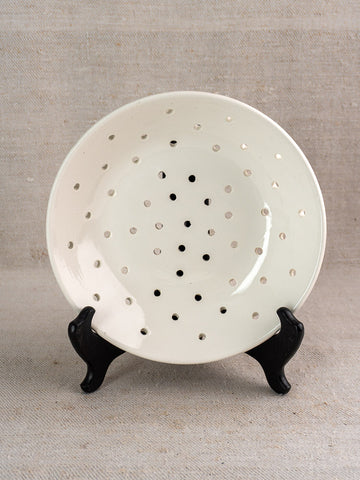 Antique French Digoin Sarreguemines berry strainer in white stoneware on black stand, beige textured fabric backdrop.