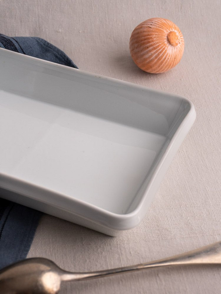 Vintage French white porcelain baking dish on light fabric with silver spoon, blue napkin, and orange seashell.