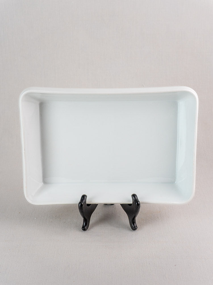 Vintage French white porcelain rectangular baking dish on black stand, neutral light gray background.