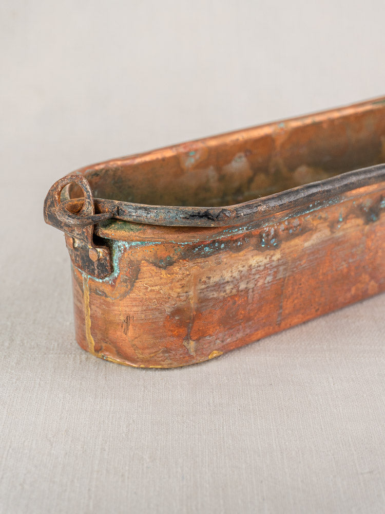 Antique French copper poissonniere fish kettle with patina, tarnish, rounded edge, and looped handle on light fabric background.