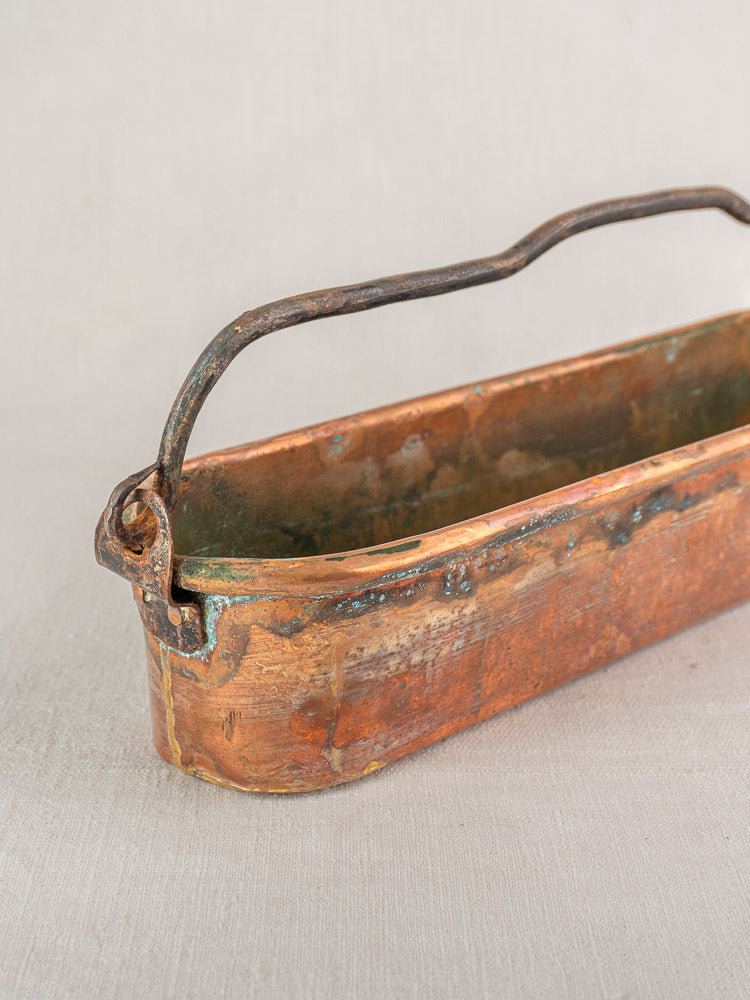 Antique French copper poissonniere with green oxidation, dual curved iron handles, on a light textured surface.