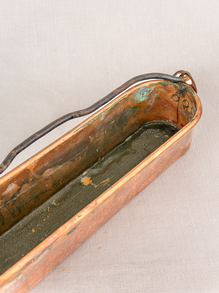 Antique French copper fish kettle with dark metal handle and worn interior, displayed on beige fabric.