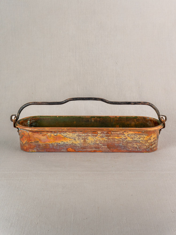 Antique French copper poissonniere with weathered patina and dark curved handle on a light gray background.