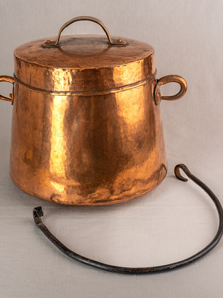 Antique French copper lidded cauldron with side handles and black double-hooked metal tool on light surface.
