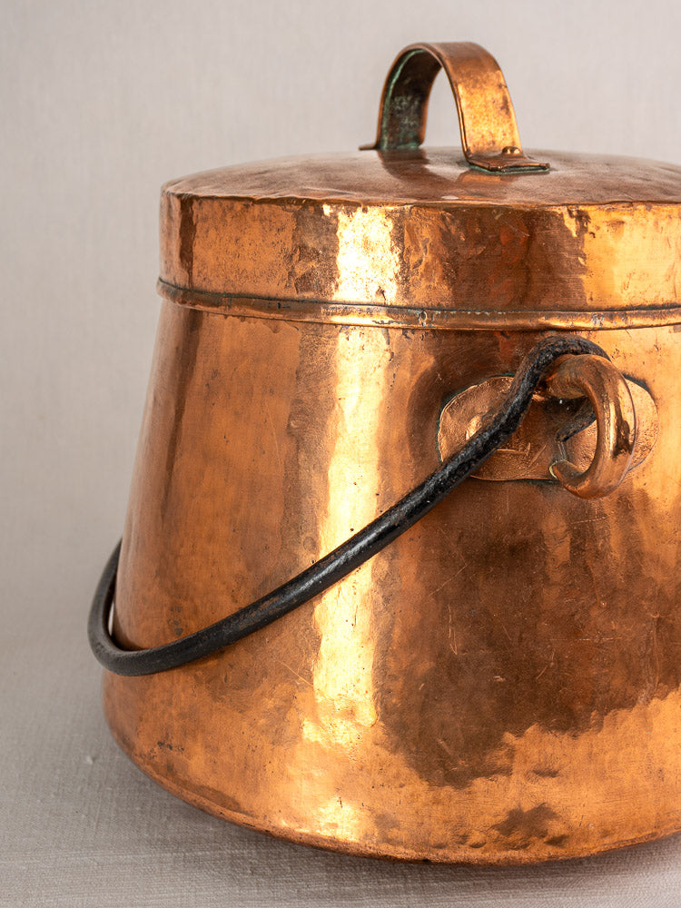 Antique French hand-hammered copper cauldron with curved top handle and black side handle on neutral background.