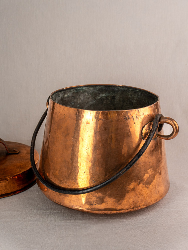 Antique French copper cauldron with hand-hammered texture and dark handle, matching lid beside on light surface.