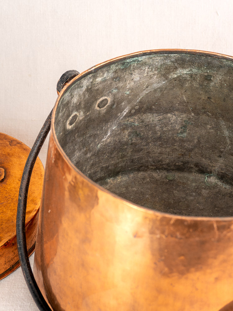 Antique French hand-hammered copper cauldron with metal handle and lid, shown on a light surface.