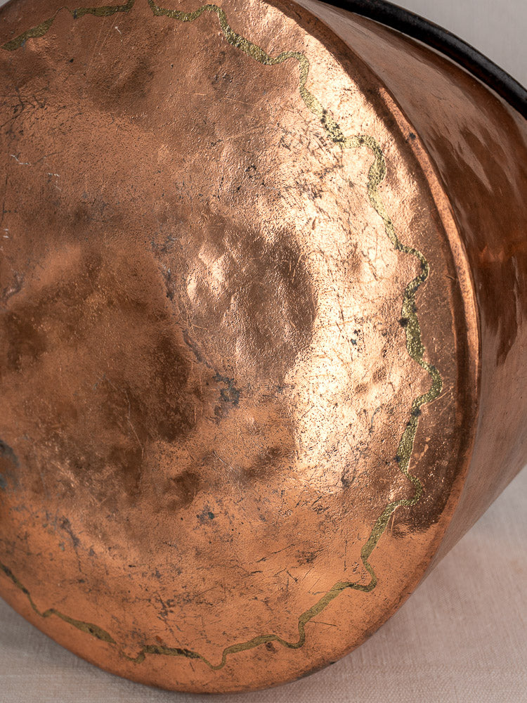 Antique French hand-hammered copper lidded cauldron with scalloped seam, oxidation, and scratches on light fabric.